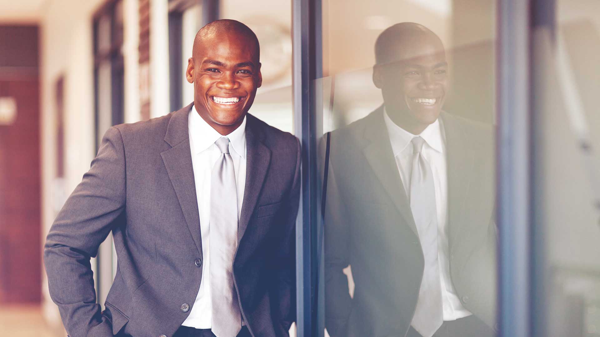 Professional man in a suit leaning up against a window smiling