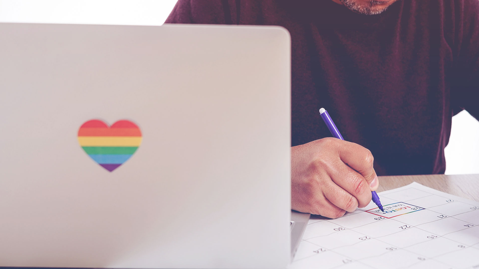 unidentifed man sitting at desk making a note about Pride on his calendar. Computer has a heart in Rainbow colors for Pride. 